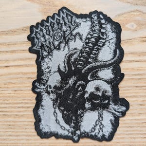 Pode incluir: Patch bordado em preto e branco com um crânio com chifres e detalhes intrincados. O patch tem um tecido texturizado e uma borda branca.
