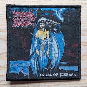 May include: Black square patch featuring the band name "MORBID ANGEL" in red, stylized lettering. The patch depicts a figure in blue robes with a pale complexion, with the text "ANGEL OF DISEASE" below the image. The patch is bordered with black.