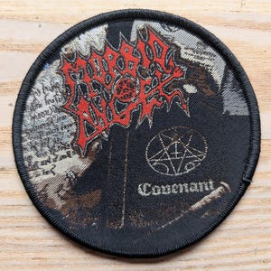May include: Circular black patch featuring a red and black logo, a pentagram, and the word "Covenant". The patch has a black border and is designed for application on clothing or bags. The logo is in a stylized font.