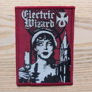 May include: A rectangular patch with a red background and a red border. The patch features a black and white illustration of a woman with a candle and a building. The words "Electric Wizard" are printed in silver.