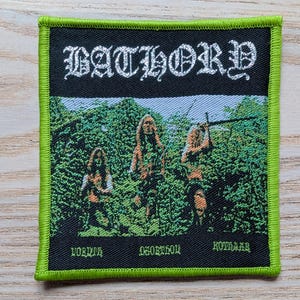 May include: Square patch with a green border and a black center featuring the band name "BATHORY" in white gothic lettering. Below the band name is an image of three figures in a forest. The bottom of the patch has text.
