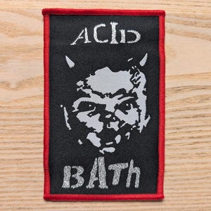 Peut inclure: Écusson rectangulaire noir avec une bordure rouge. L'écusson porte les mots "ACID BATH" en blanc, avec une illustration blanche d'un visage avec des cornes. L'écusson est probablement destiné à une veste ou un sac.