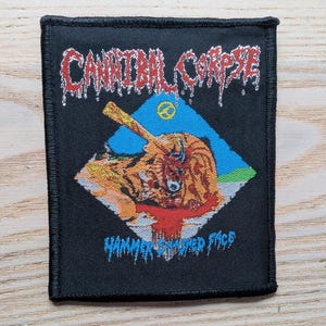Könnte beinhalten: Schwarzer, quadratischer Aufnäher mit dem Bandnamen "Cannibal Corpse" in roter, tropfender Schrift. Der Aufnäher zeigt eine Grafik einer gewalttätigen Szene mit dem Text "Hammer Smashed Face" darunter. Ein Friedenszeichen ist sichtbar.