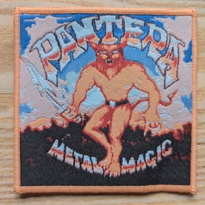 May include: Square patch with an orange border featuring the word "PANTERA" in white, above an illustration of a red demon holding a sword. The words "METAL MAGIC" are below the demon. The background is blue and peach.