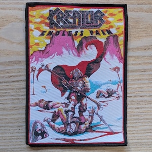 May include: Embroidered patch with the band name "Kreator" and the album title "Endless Pain." The artwork shows a warrior with a weapon, surrounded by fallen figures, against a mountainous backdrop. The patch has a black border.
