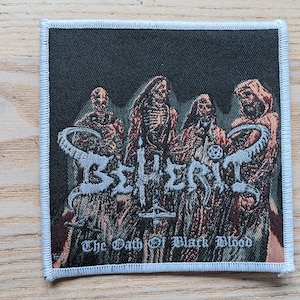 Könnte beinhalten: Quadratischer Aufnäher mit schwarzem Hintergrund, der ein Bandlogo und Figuren in braunen Roben zeigt. Der Text "BEHERIT" ist in Weiß, darunter "The Oath of Black Blood". Der Aufnäher hat einen weißen Rand.