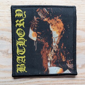 May include: A square black patch with the word "BATHORY" in yellow gothic lettering. The patch features a detailed illustration in shades of brown, orange, and white. The patch has a black border.