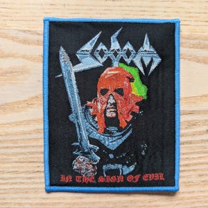 Può includere: Toppa nera con bordo blu, raffigurante una figura mascherata di rosso che tiene una spada. Il nome della band "Sodom" è sopra e il testo "In the Sign of Evil" sotto. Il design include accenti verdi e bianchi.