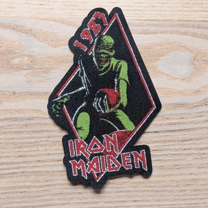 Könnte beinhalten: Gestickter Aufnäher mit einer grünen, zombieartigen Figur, die ein rotes Objekt hält, vor einem schwarzen Diamanten. Der Aufnäher enthält den Text "1987" und "Iron Maiden" in Rot und Weiß. Der Aufnäher befindet sich auf einer Holzoberfläche.