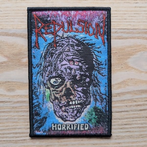 May include: A rectangular patch with a black border features a zombie-like face with the word "REPULSION" in red above it. The word "HORRIFIED" is printed in white at the bottom. The background is blue and purple.