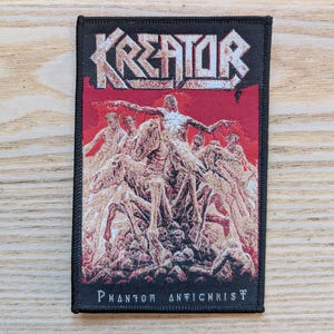 Könnte beinhalten: Schwarzer rechteckiger Aufnäher mit dem Wort "KREATOR" in weißer, stilisierter Schrift über einem roten Hintergrund. Das Bild zeigt skelettartige Figuren. Der Text "PHANTOM ANTICHRIST" befindet sich unten.