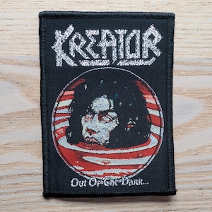 Peut inclure: Écusson rectangulaire noir avec le nom du groupe "KREATOR" en lettres blanches stylisées. En dessous, un graphisme circulaire d'un visage pâle sur fond rayé rouge et blanc. Le texte "Out Of The Dark..." est en dessous.