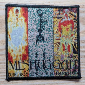 Op de afbeelding: Vierkantige patch met een zwarte rand met een ontwerp met drie verticale panelen. De panelen tonen abstracte figuren in rode, gele en groene tinten. De bandnaam "MESHUGGAH" en de woorden "DESTROY ERASE IMPROVE" zijn in het zwart gedrukt.