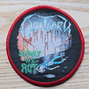 May include: Round black patch with a red border. The patch features the word "OBITUARY" in blue and white, with red drips. Below, the words "SLOWLY WE ROT" are in green. A blue figure is at the bottom.