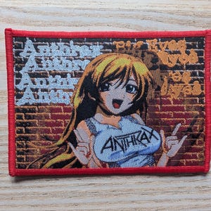 May include: A rectangular embroidered patch with a red border featuring an anime-style character with long blonde hair against a brick wall background. The text Anthrax is repeated in light blue on the left and in black on the character's shirt.