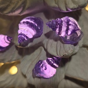 May include: Four translucent purple seashell-shaped ornaments are attached to a white, textured, conical structure. The seashells have a spiral design and emit a soft, glowing light, contrasting with the matte white surface of the tree-like object.