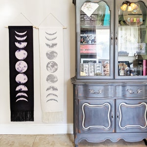 May include: Two black and white moon phase tapestries hanging on a white wall next to a gray painted antique cabinet with glass doors. The cabinet is decorated with a variety of items, including a small plant, a skull, and a sign that reads "CARDS".