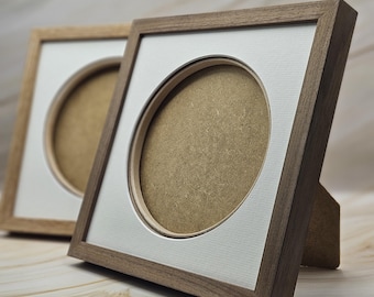 Peut inclure: Deux cadres photo carrés en bois avec une finition brun foncé. Chaque cadre présente un passe-partout blanc avec une découpe circulaire au centre, révélant un fond de couleur beige. Les cadres sont positionnés en angle sur une surface.