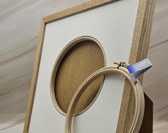 Peut inclure: Un cercle à broder en bois et un cadre photo en chêne clair avec une ouverture ovale. Le cadre a un passe-partout blanc et un dos marron. Une petite étiquette est attachée au cercle à broder. Le cadre est posé contre un support en bois.