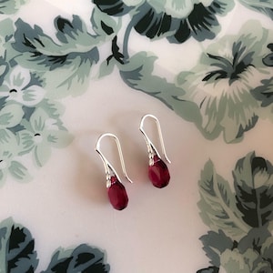 May include: A pair of silver-toned drop earrings featuring faceted, teardrop-shaped red crystals. Each earring has a curved metal hook design that holds the vibrant red stone at the base, creating a simple and elegant dangling jewellery piece.