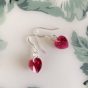 May include: A pair of heart-shaped earrings in a vivid red colour. The earrings have a faceted design, reflecting light. The hooks are made of silver-coloured metal, creating a delicate and romantic accessory.