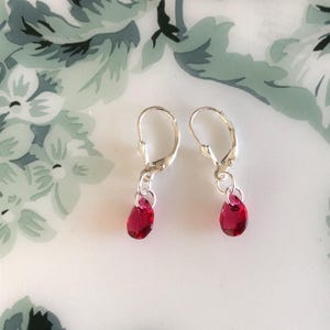 May include: A pair of silver leverback earrings with teardrop-shaped, deep red crystal pendants. The earrings are displayed on a white surface with a floral pattern.