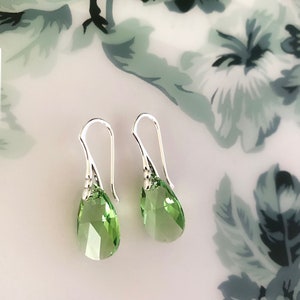 May include: A pair of silver drop earrings featuring faceted, pear-shaped, light green crystals. The earrings are presented on a white surface with a floral pattern in green and grey.