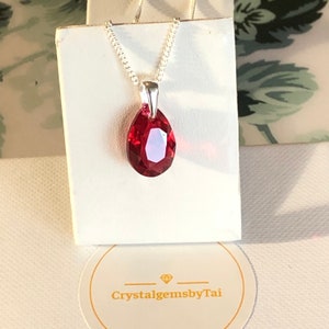 May include: A silver chain necklace with a teardrop-shaped red crystal pendant. The pendant is faceted and has a high shine. The necklace is displayed on a white card with the text "CrystalgemsbyTai".