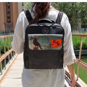 May include: A black leather backpack with a colourful graphic of a crow singing into a microphone with the word "GAK" in a speech bubble. The backpack has a front pocket with a zip.