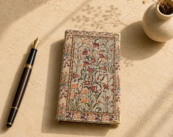 May include: A rectangular notebook with a woven fabric cover featuring a multi-colored floral pattern in shades of red, orange, and green, accented with gold-toned metal corner protectors, positioned next to a black fountain pen with gold trim.