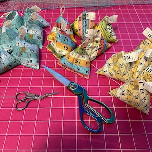 May include: A selection of small, triangular fabric pouches patterned with tape measures, and two pairs of scissors. The pouches feature teal, yellow, and orange tape measure designs. The scissors have iridescent handles. The measurements are in cm.