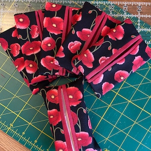 May include: A selection of fabric tissue holders and cases. The holders are black with a red poppy floral pattern. The edges are trimmed with a burgundy ribbon. The tissue holders are arranged on a green cutting mat.