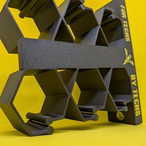 Puede incluir: Un estante de almacenamiento negro impreso en 3D con una estructura de rejilla en forma de panal. El panel lateral presenta el texto amarillo "FOR TECHS BY TECHS" acompañado de un icono de abeja. La unidad tiene múltiples compartimentos abiertos.