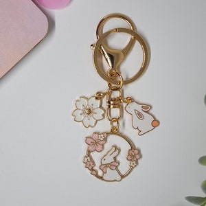 May include: Gold-tone keychain with three charms: a white flower, a white bunny, and a bunny in a pink flower wreath. The charms are accented with pink and gold details. The keychain has a lobster clasp and a split ring.