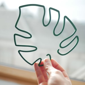 Könnte beinhalten: Ein grünes Drahtgeflecht in Form eines Monstera-Blattes. Das Blatt wird in der Hand einer Person gehalten.