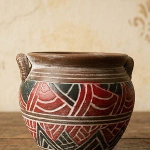 May include: A round brown ceramic pot featuring two small side handles and a wide rim. The exterior surface is decorated with a repeating geometric pattern in deep red, black, and white, characterized by curved lines and triangular shapes.
