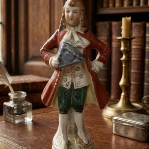 May include: A porcelain figurine of a person wearing an 18th-century style red coat, white cravat, and green breeches. The figure has brown hair and holds a triangular gray object in the right hand, standing on a decorative circular base.
