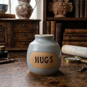 May include: A small, round, light blue ceramic jar with a speckled finish and a wide opening. The front features a textured, tan-colored oval patch with the word HUGS embossed in dark, bold, capital letters across the center of the surface.