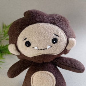 May include: A handmade plush toy in the shape of a cartoon Bigfoot. The toy has a brown body and head with a tan face, ears, and belly. It features black eyes, a small smile with two white teeth, and is designed to resemble the mythical creature.