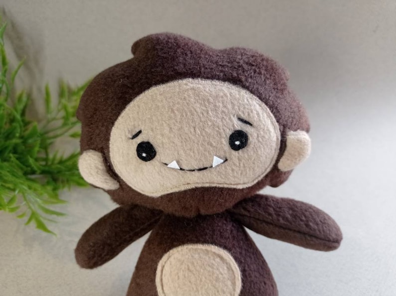 May include: A handmade plush toy in the shape of a cartoon Bigfoot. The toy has a brown body and head with a tan face, ears, and belly. It features black eyes, a small smile with two white teeth, and is designed to resemble the mythical creature.