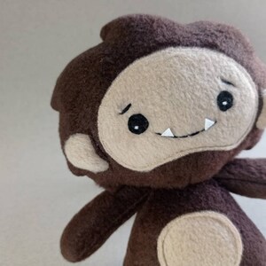 May include: A handmade plush toy in the shape of a cartoonish Bigfoot. The toy is primarily brown with a tan face, arms, and feet. It has black eyes, a small mouth with white teeth, and a slightly furrowed brow.