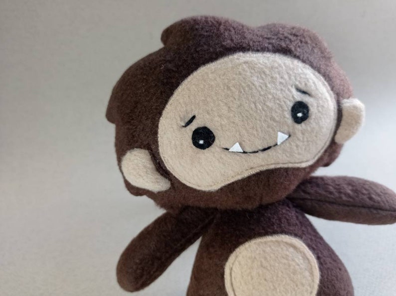 May include: A handmade plush toy in the shape of a cartoonish Bigfoot. The toy is primarily brown with a tan face, arms, and feet. It has black eyes, a small mouth with white teeth, and a slightly furrowed brow.