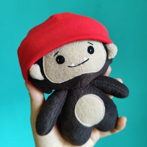 May include: A handmade plush toy monkey with a red cap. The monkey has a tan face, dark brown body, and a white belly. The toy has black eyes and a small smile. The background is a solid teal color.