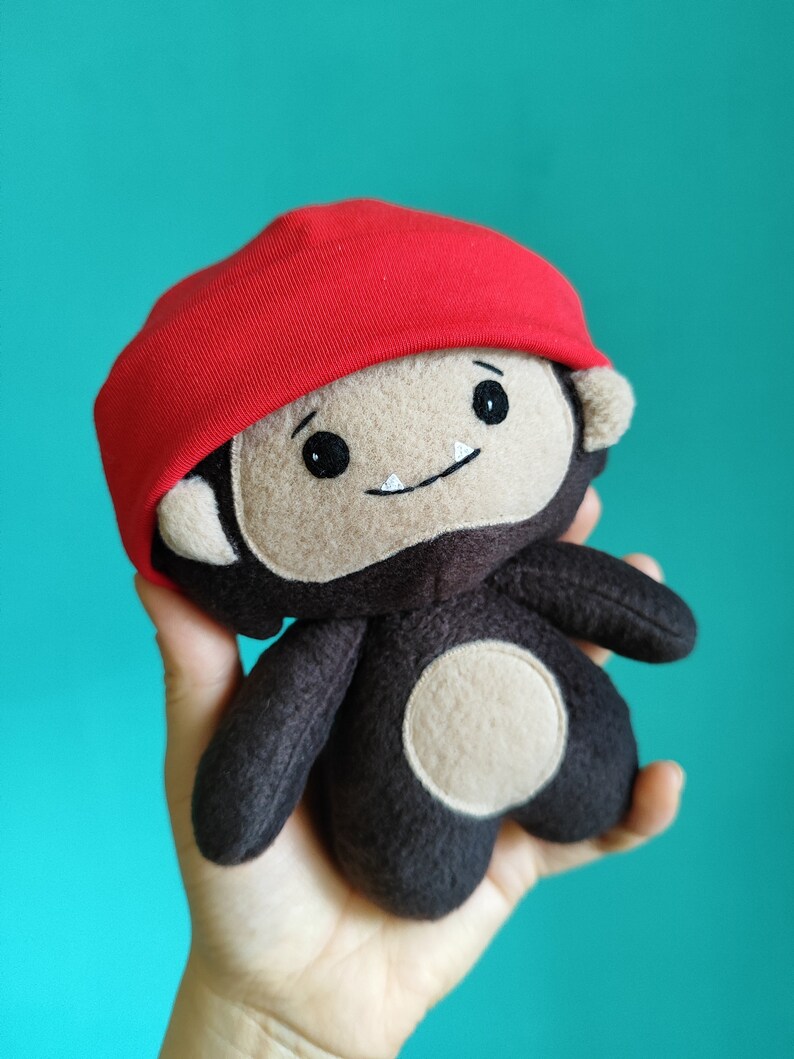 May include: A handmade plush toy monkey with a red cap. The monkey has a tan face, dark brown body, and a white belly. The toy has black eyes and a small smile. The background is a solid teal color.