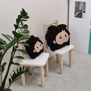 May include: Two white wooden chairs with brown plush cushions featuring cartoon faces. The chairs are on a light gray carpet in a room with a white wall and a green plant.