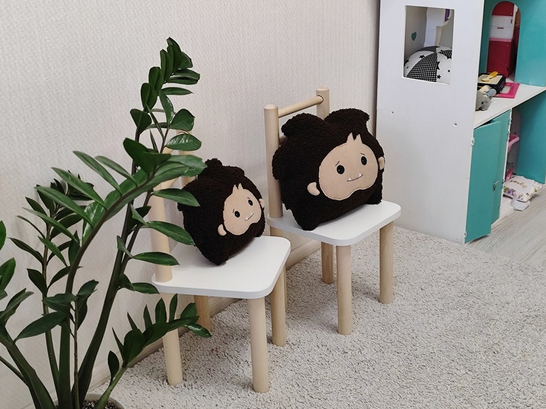 May include: Two white wooden chairs with brown plush cushions featuring cartoon faces. The chairs are on a light gray carpet in a room with a white wall and a green plant.
