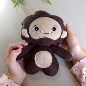May include: A handmade plush toy in the shape of a cartoon bigfoot. The toy is primarily brown with a tan face, ears, and belly. It has a smiling expression with two white teeth. The toy is being held by a person.