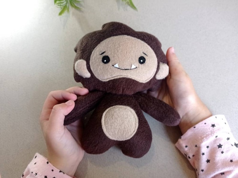 May include: A handmade plush toy in the shape of a cartoon bigfoot. The toy is primarily brown with a tan face, ears, and belly. It has a smiling expression with two white teeth. The toy is being held by a person.