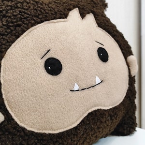 May include: A brown plush pillow with a beige face, black eyes, and white teeth. The pillow is shaped like a monster.