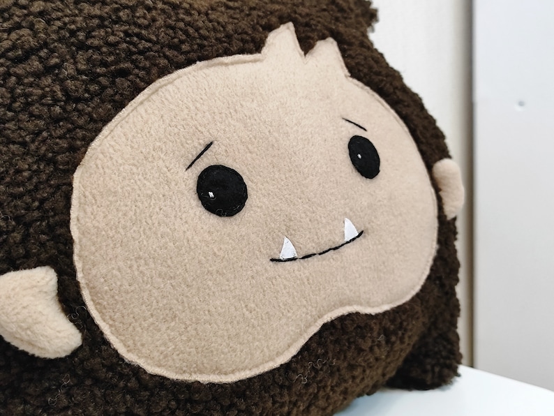 May include: A brown plush pillow with a beige face, black eyes, and white teeth. The pillow is shaped like a monster.